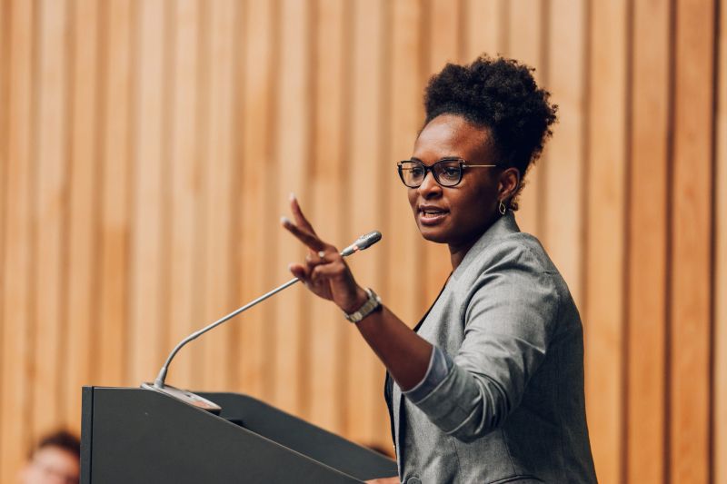 Black woman speaking at a podium, gesturing with her hand while delivering a speech during a professional conference