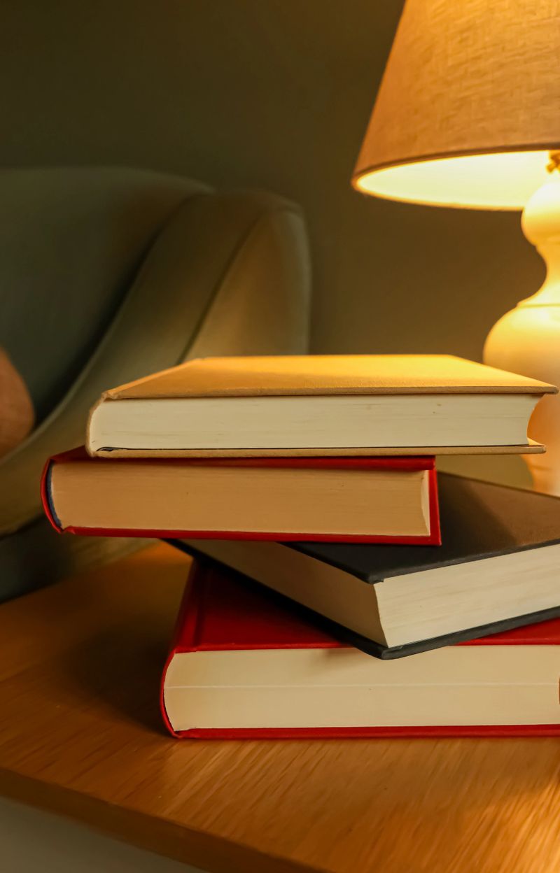 Stacked hardcover books resting on a wooden table beside an illuminated lamp, symbolizing education, knowledge, and quiet relaxation