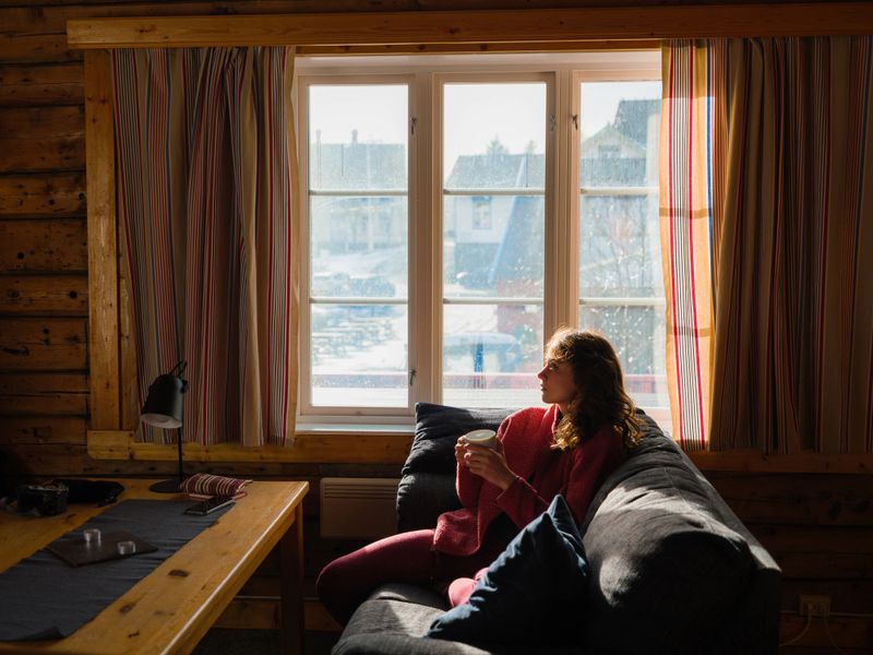 Woman drinking coffee and relaxing on the sofa in a cozy wooden  cabin on Lofoten islands in winter