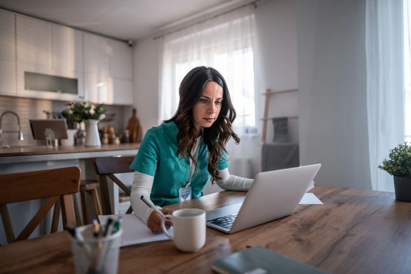 Female doctor or nurse in teal scrubs working remotely, focused on a laptop and taking notes at a wooden table in her modern home office, representing telehealth and distance education