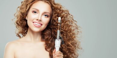 A smiling woman with voluminous spiral curls holds a curling iron.