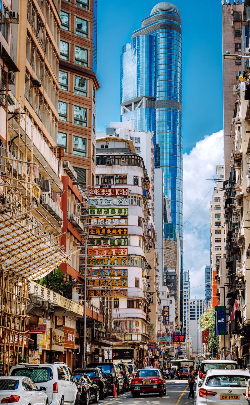 Bustling Hong Kong street framed by aging midrise buildings and a towering glass skyscraper. Colorful signs and dense traffic illustrate the city’s vibrant contrast between old and new.