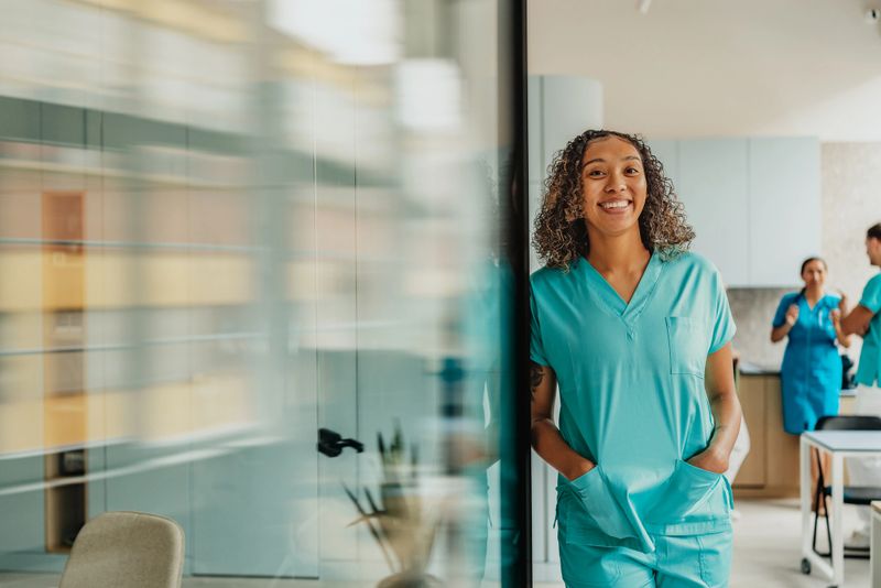 In a bright and modern medical clinic, a healthcare worker in scrubs smiles confidently while colleagues engage in discussion nearby. The atmosphere radiates teamwork and support.