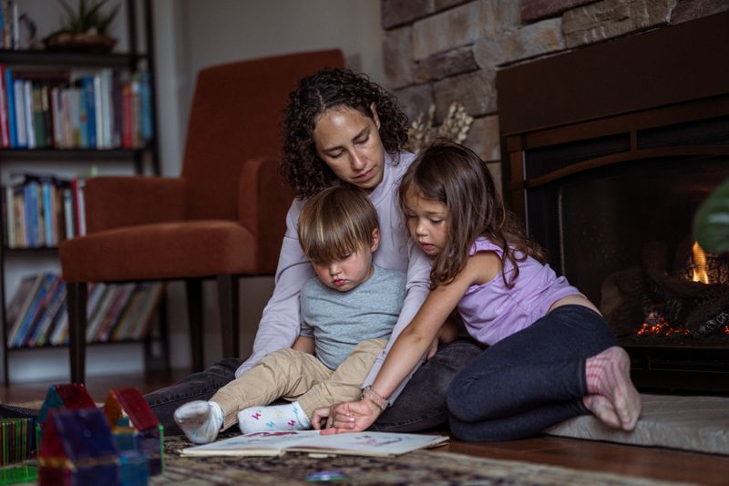 A multiracial woman of Pacific Islander and Asian descent sits on the living room floor in front of the fireplace with her two-year-old son and five-year-old daughter and reads a book to them while enjoying a cozy and relaxing day together at home.