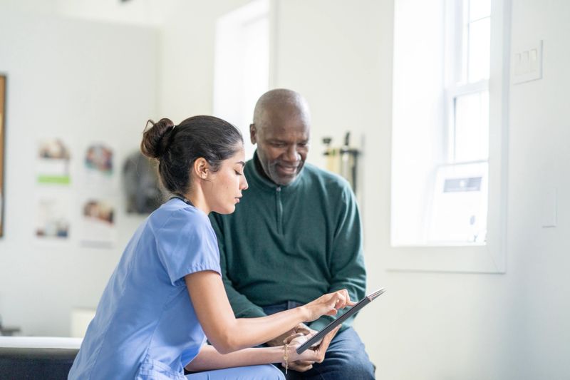 A compassionate nurse in blue scrubs sits with an elderly Black man, reviewing care information on a tablet in a bright clinic. The scene highlights diverse healthcare teamwork and clear patient communication.