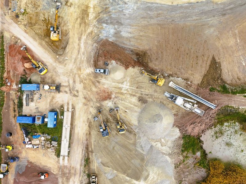 Top-down drone view of an active construction site featuring heavy machinery, including an excavator and bulldozer, surrounded by blue temporary site offices and storage containers. The image captures the organized layout, construction materials, and earthwork area, ideal for illustrating industrial development, infrastructure projects, or engineering concepts.