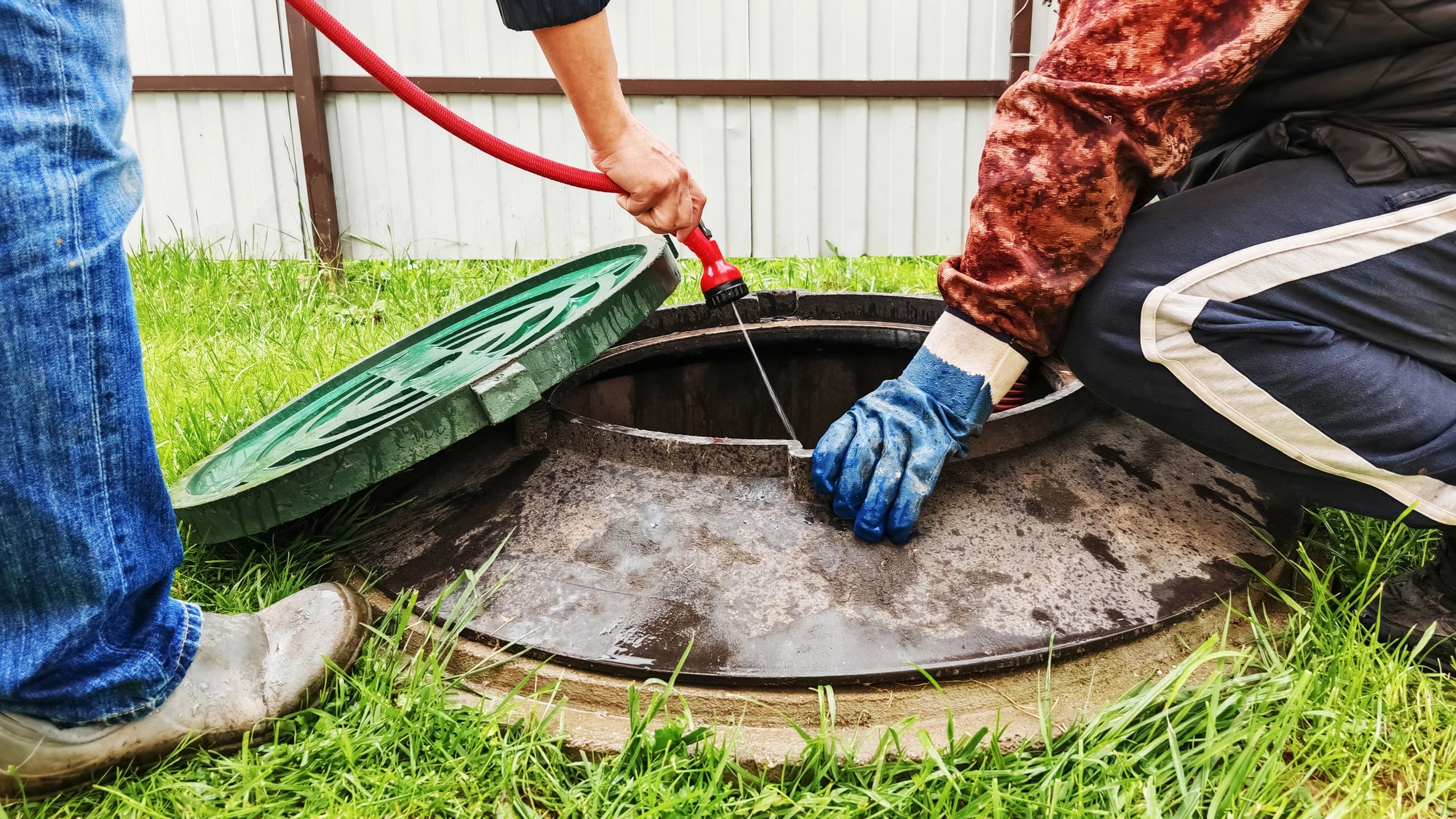 Two men cleaning a manhole with a hose and gloves in a grassy area.