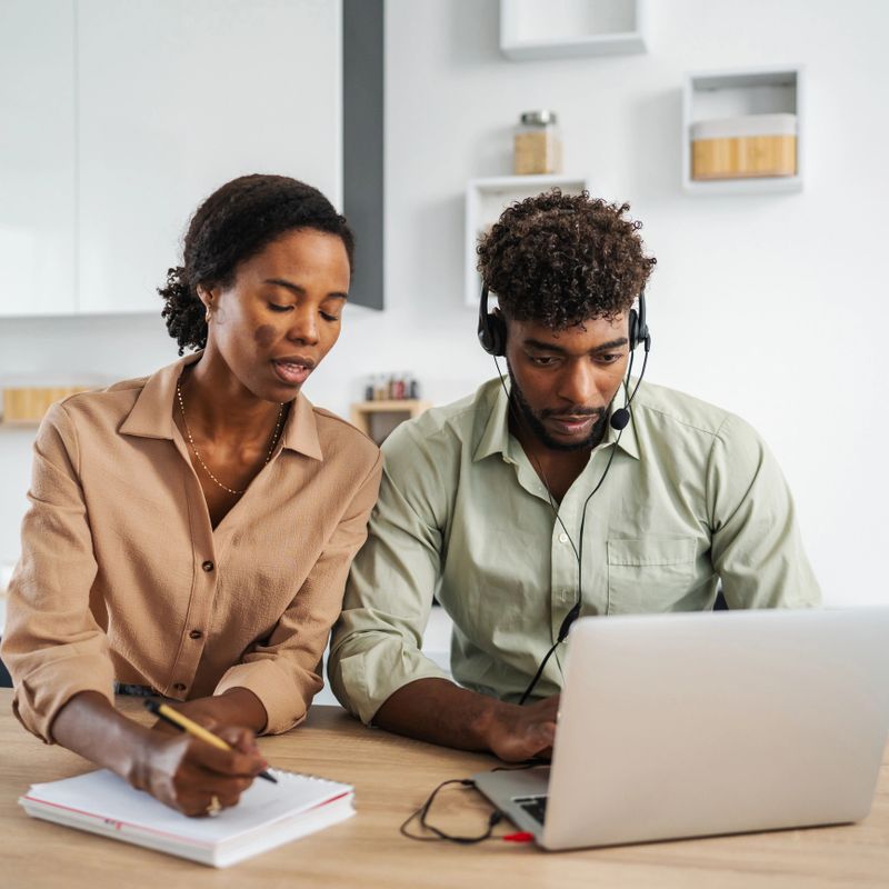 Black man wearing a headset and using a laptop with a black woman taking notes in a notebook, both collaborating on a project from their modern kitchen