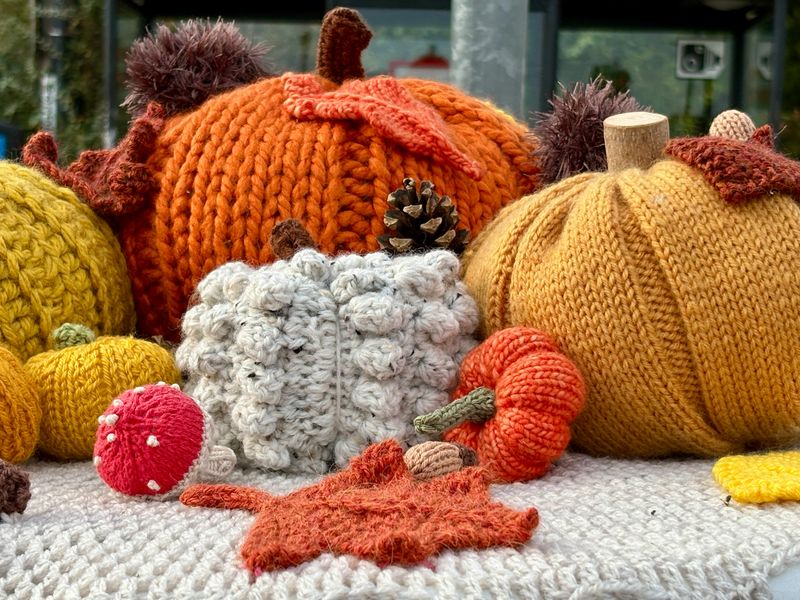 A cozy autumn display featuring hand-knitted pumpkins, leaves, mushrooms, and acorns in warm shades of orange, yellow, white, and brown, beautifully crafted with textured yarn and charming detail.