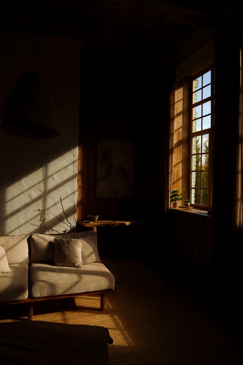 Sunlight fills a cozy room with wooden elements casting shadows on a sofa. A tranquil atmosphere is created with plants near the window perfect for relaxation in the evening.