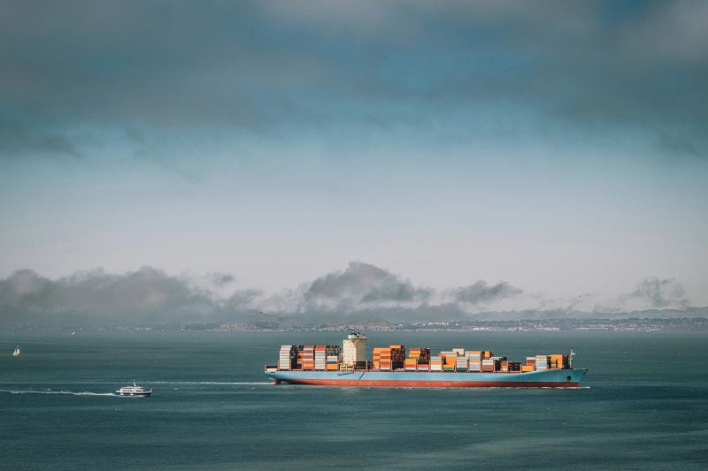 A large blue and orange container ship carrying steal shipping crates across the blue ocean, followed by a smaller boat during the day.