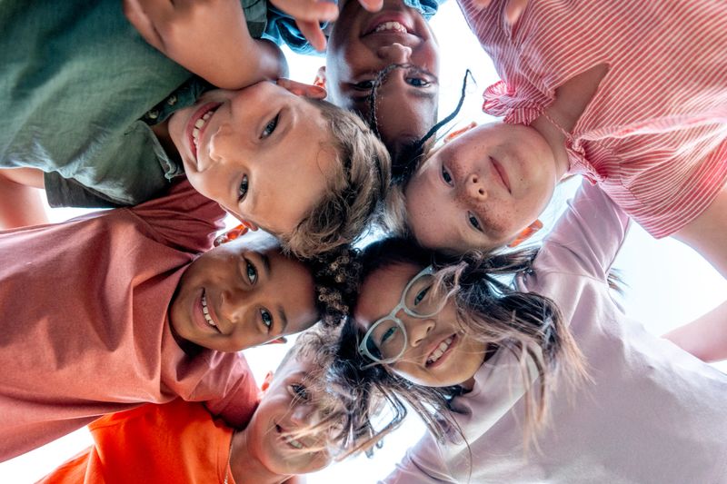 A diverse group of smiling children form a circle and look down at the camera, capturing friendship, teamwork, and carefree summer fun in a bright outdoor setting.