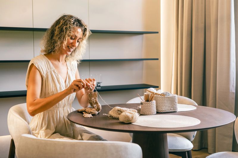 Happy woman sitting at a round table, crocheting with brown yarn, surrounded by crochet tools and yarn baskets in a softly lit, cozy modern interior with natural daylight.