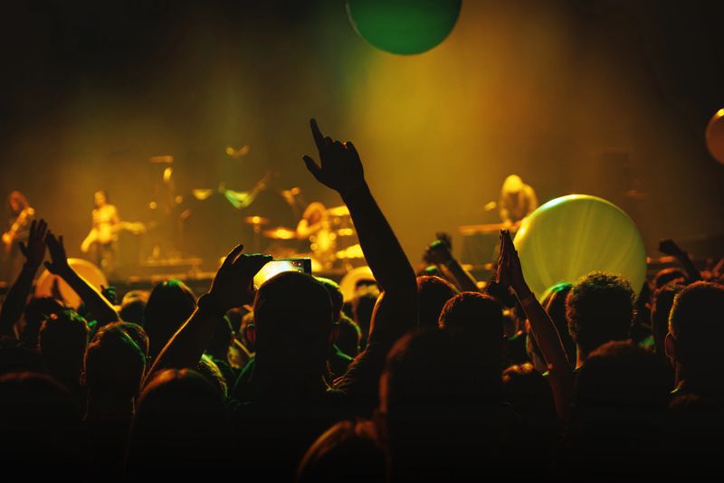 Audience members raise hands during a concert while green and yellow lights glow on stage. The picture captures emotion, enthusiasm, and collective rhythm of live music events