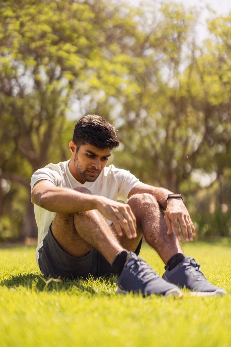 A lone man rests on the bright green grass in a sunny park, knees up and arms draped over them. He looks inward, wearing a white t‑shirt, dark shorts, and athletic shoes. The scene conveys calm, reflection, and outdoor fitness enjoyed in nature.