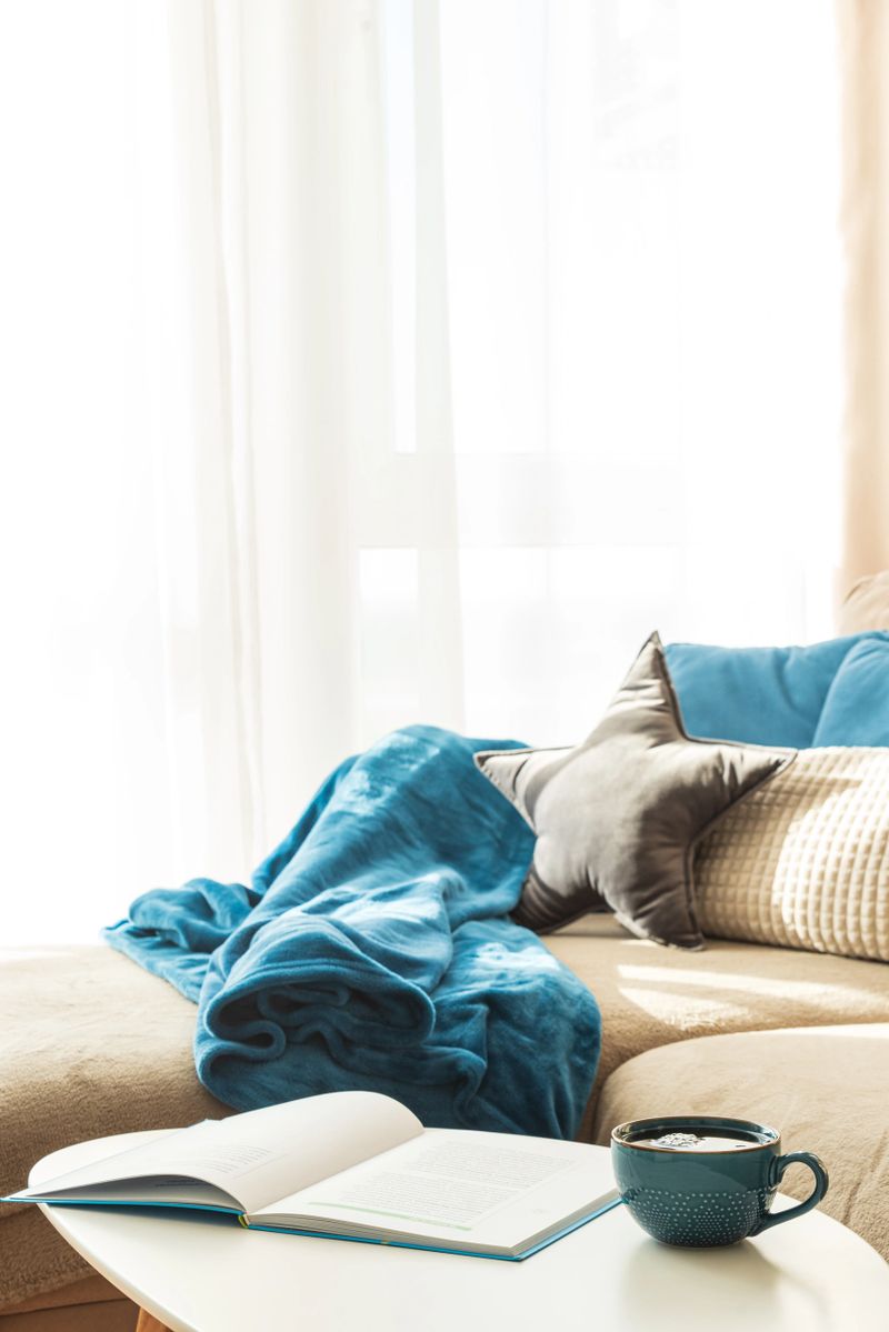 Cozy Living Room with mug and book on Coffee Table, Cushions, Blanket and Sunlight on Sofa