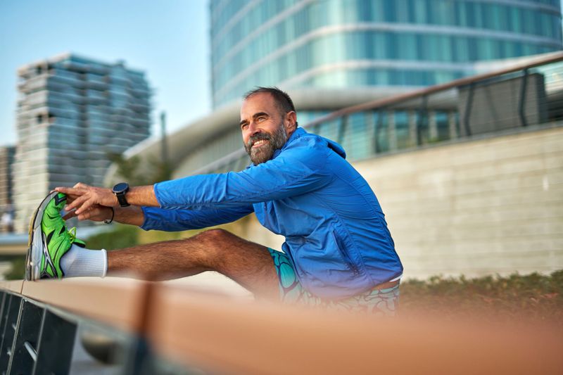 Mature bearded man smiling while stretching to touch his toes before a run in a sunny modern city, dressed in sportswear and preparing for an outdoor workout and jogging session