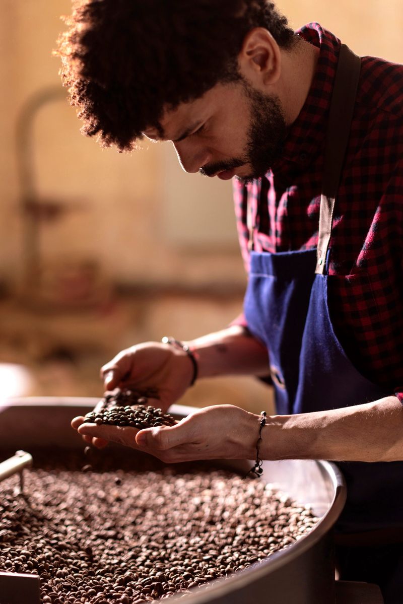 A focused barista in a blue apron examines dark roasted coffee beans in a large roasting tray; Warm lighting and a craft-focused kitchen setting convey artisanal coffee production and attention to detail
