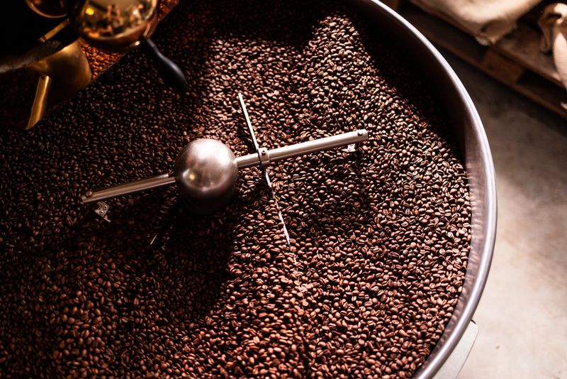 Rich coffee roasting scene inside a metal drum shows glossy beans and a stainless steel stirrer under warm light, capturing the energy and craft of small-batch production