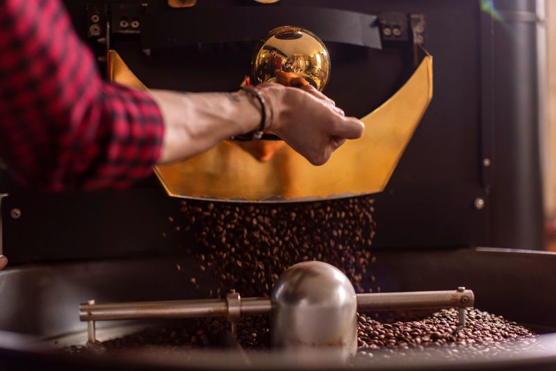 Close-up of a baristaâs hands turning a lever on a gold-toned coffee roaster, as beans cascade into a tray; Warm, aromatic process conveying craft, focus, and cafe atmosphere