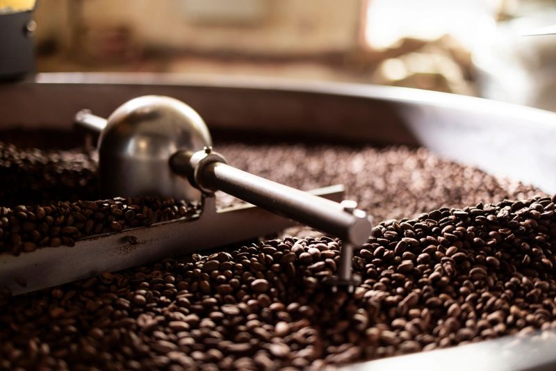 Close-up of a coffee roasting process featuring a metal scoop over a bed of glossy brown beans in a production facility; Warm lighting and industrial equipment convey energy and craftsmanship