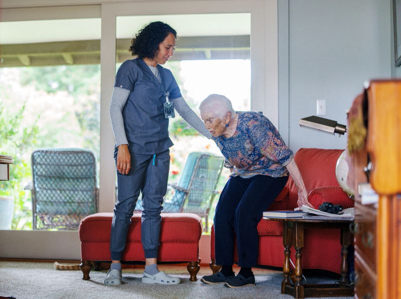 A home health nurse helps an elderly woman stand up from the chair during a home visit, offering gentle support and care.