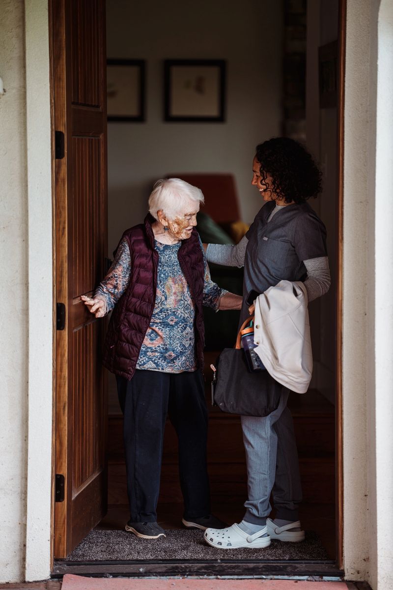 An Eurasian woman who works as a home health nurse cheerfully greets and chats with an elderly patient when arriving at the woman's home at the start of a work shift.