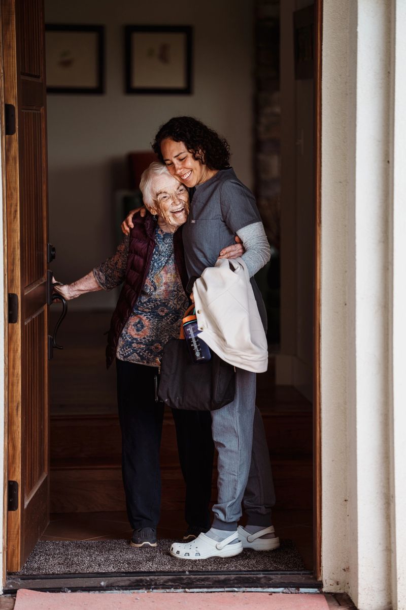 A smiling elderly woman opens the front door to her home and greets her female home health nurse with a hug.