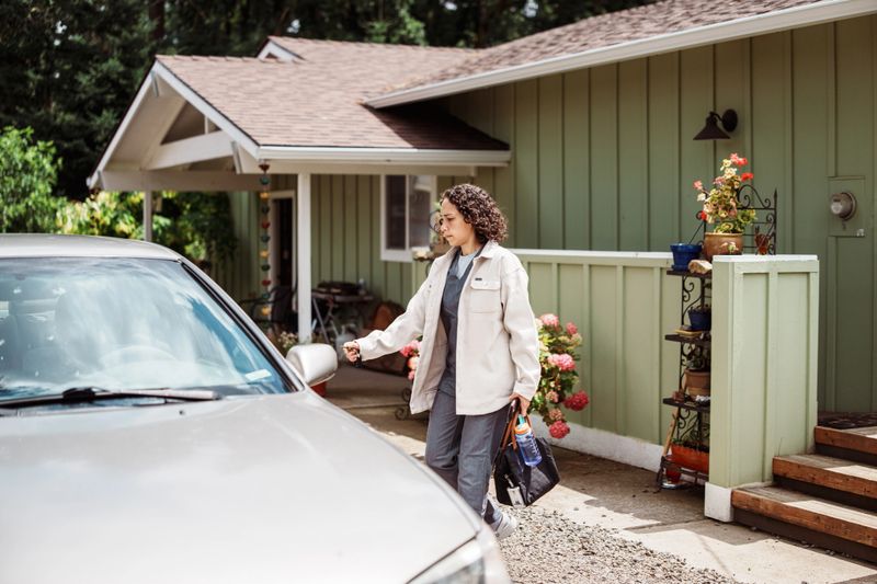 A Eurasian woman dressed in scrubs and a cream jacket prepares to leave for work, unlocking her car parked in the driveway outside her home.