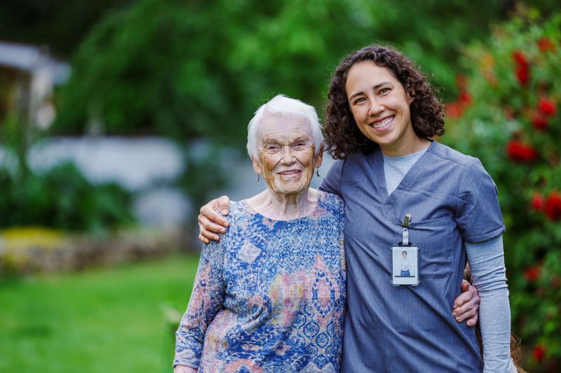 Portrait of an Eurasian female home health nurse and her elderly female patient affectionately embracing and smiling directly at the camera while enjoying time together outdoors in the patient's rural and beautifully landscape back yard.
