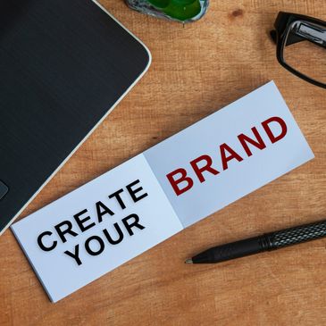 A note on a desk says 'CREATE YOUR BRAND' with a laptop, pen, and glasses nearby.