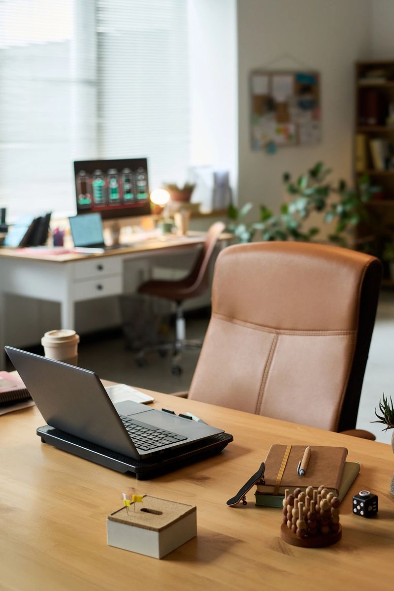 Modern office workspace featuring open laptop, notebooks, pen, coffee cup, and desk accessories on wooden table with desktop computer and plants visible in background