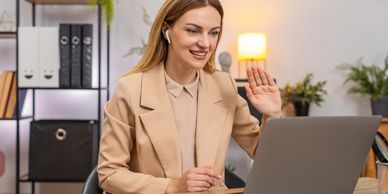 A woman in a beige blazer waving during a video call on her laptop.