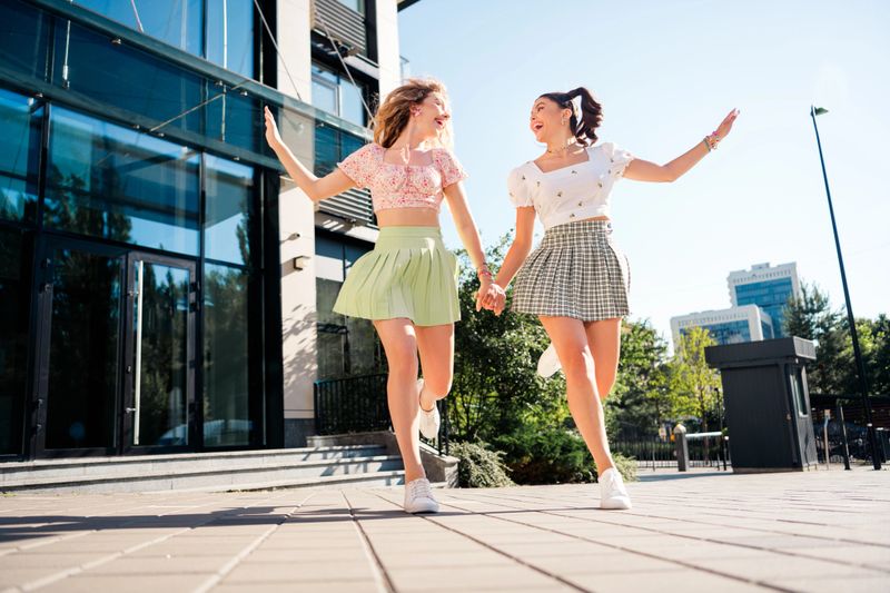 Two young women in summer outfits share a lively outdoor moment in the city, smiling and jumping together near contemporary buildings to convey friendship fashion and youthful energy in an urban setting.