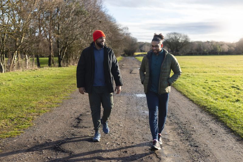 A front full-length view of two mid-adult multiracial male friends walking side by side along a dirt track on a cool day in Gosforth, England. They are wearing casual, warm outdoor clothing. The natural setting and open space convey calm, reflection, and connection. The image represents friendship, open conversation, mental health awareness, and the importance of staying active for overall well-being.Videos are available similar to this scenario.