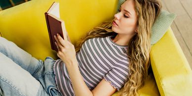 Young woman reading a book while lying on a yellow couch.