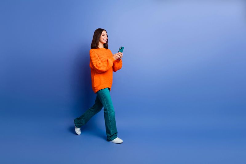 A cheerful young woman dressed in an orange knit sweater walking while using her smartphone on a blue background. Perfect representation of a happy, stylish lifestyle and technology focus.