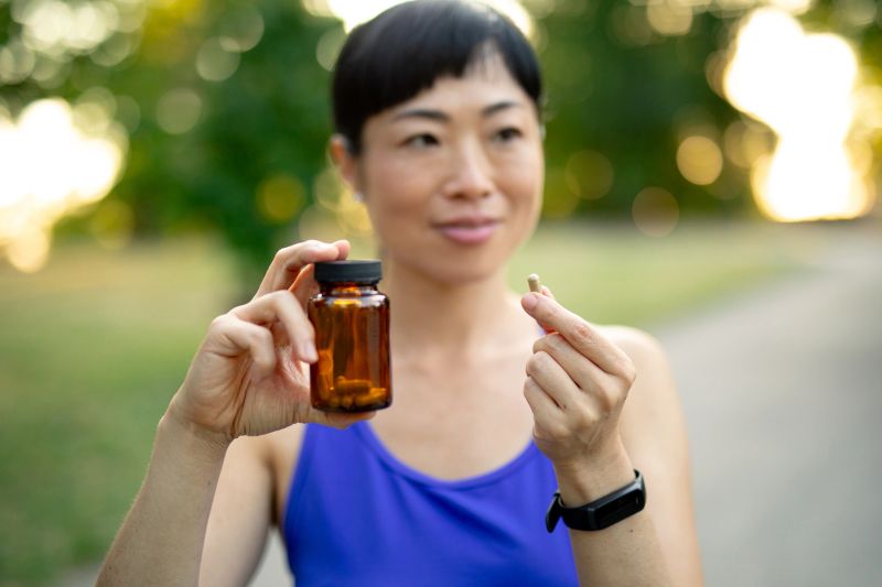 Mid-adult woman outdoors holding vitamin capsule and pill bottle, smiling and promoting daily wellness, nutrition, and active lifestyle