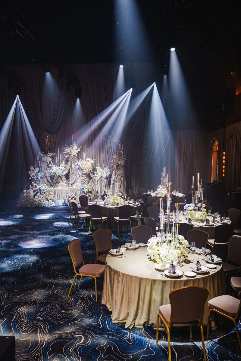 Tables are beautifully arranged in a luxurious ballroom, illuminated by focused lights. Elegant floral centerpieces enhance the sophisticated atmosphere of the venue, perfect for a celebration.