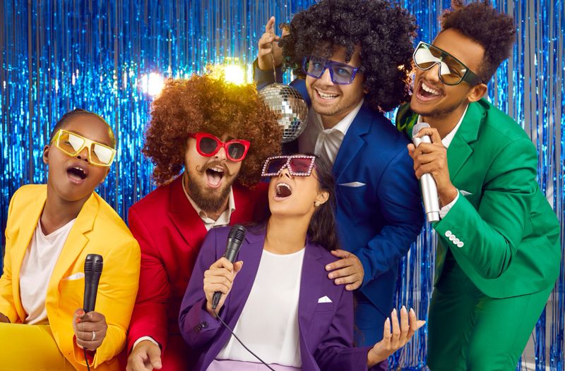 Happy multiracial friends wearing funny curly wigs and colourful disco suits singing karaoke in night club party on blue shiny background. Laughing people in sunglasses in gangnam style having a fun.