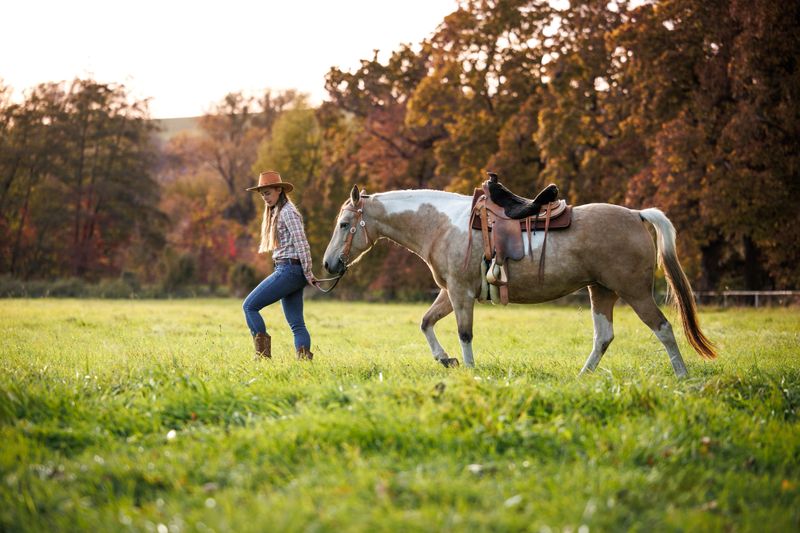 Woman in western clothing leading horse across pasture during autumn sunset. Cowgirl walking with horse, symbolizing slow living and country lifestyle