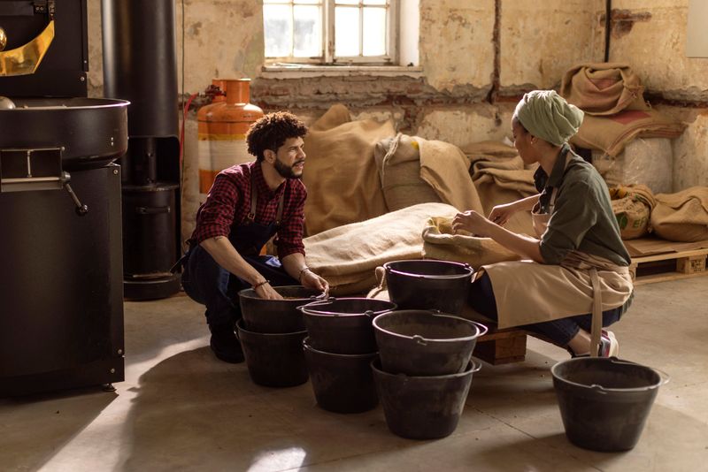 Two workers in a rustic, industrial workshop bag coffee beans beside roasting machinery and burlap sacks; They wear aprons and talk, highlighting teamwork, craft, and hands-on coffee production