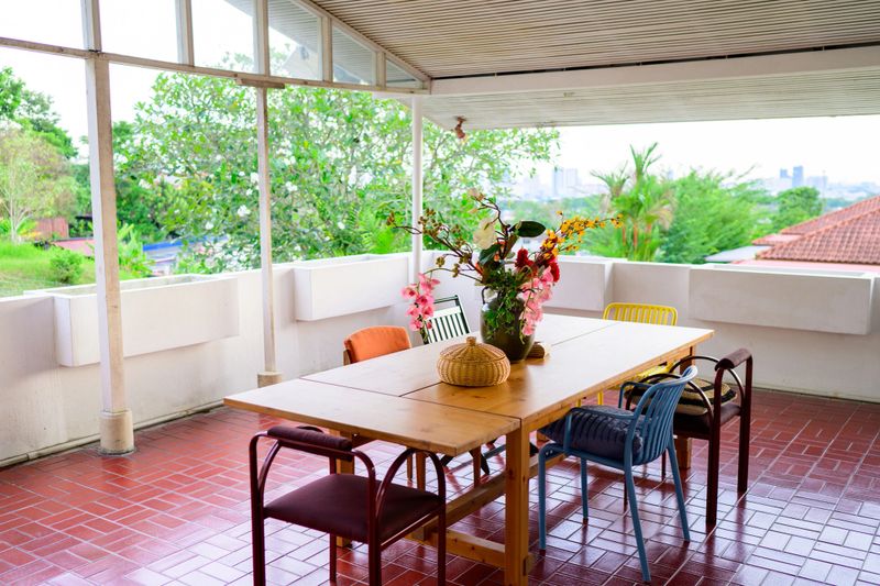 Photo of a cozy semi-outdoor dining terrace featuring a wooden table, mixed chairs, and a floral centerpiece, surrounded by lush tropical greenery.