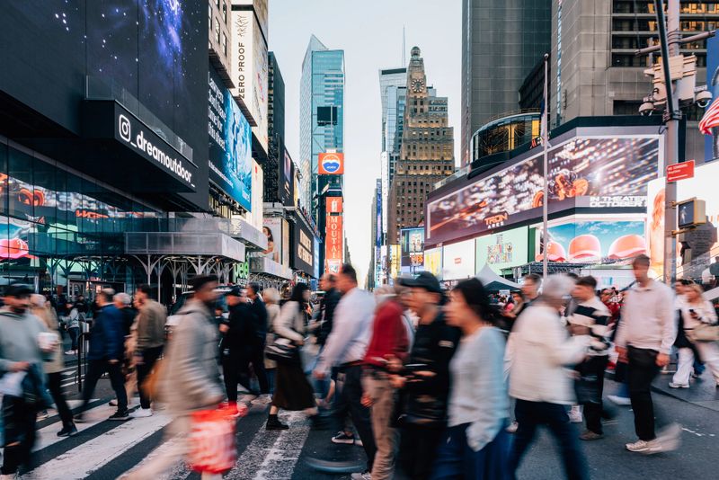A lively moment in New York City as locals and visitors move through the streets filled with signs, stores, and vibrant motion.