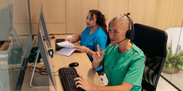 Two medical professionals in scrubs working at computers with headsets.