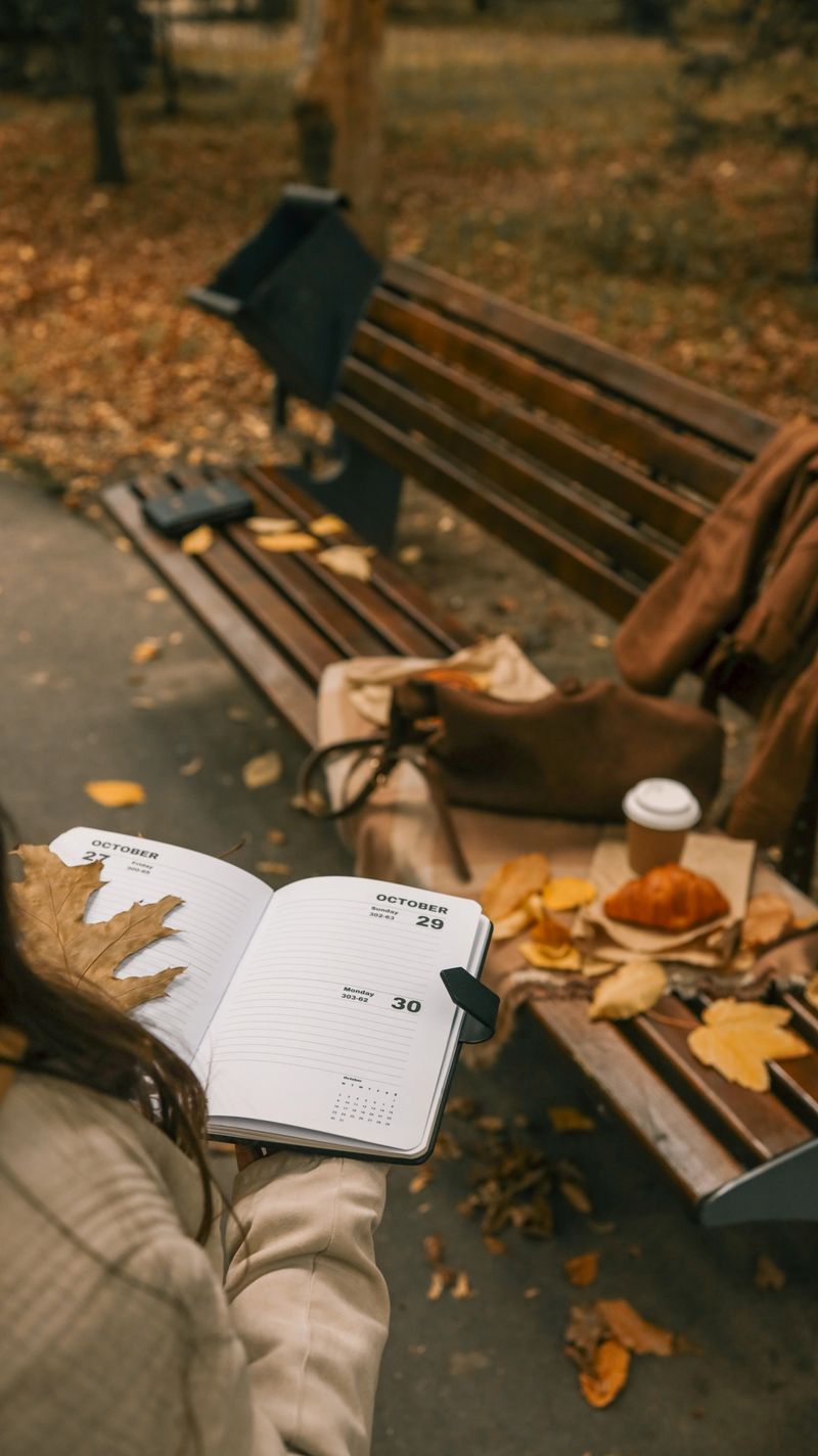 A person is holding a notebook open to October dates, with a bench, fall leaves, and a croissant in the background, creating a cozy autumn atmosphere.