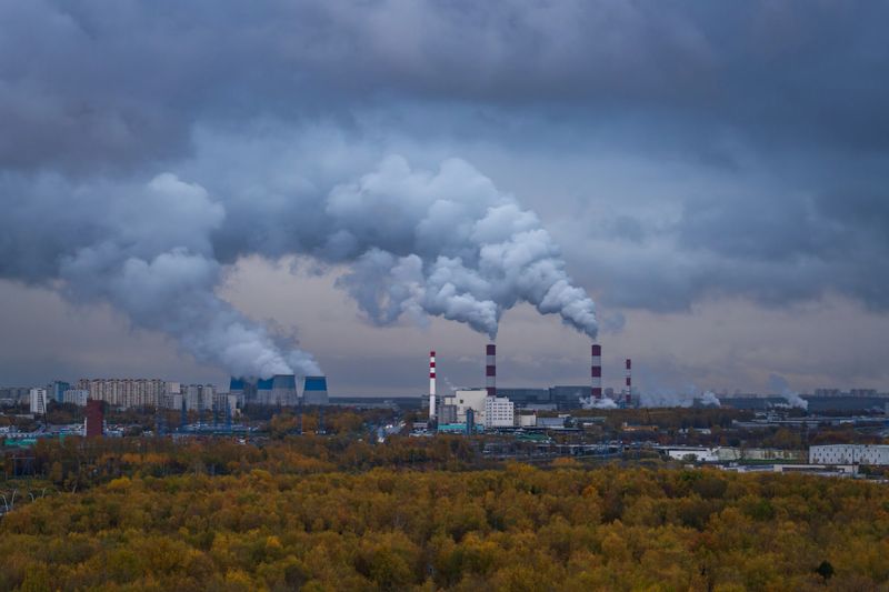 Massive plumes of smoke and steam rise from cooling towers and red‑white smokestacks across a Russian cityscape, framed by autumn forest and heavy clouds. The panoramic view emphasizes industry, environmental impact, climate, and urban air quality concerns with ample copy space.