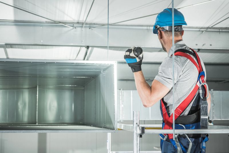 A construction HVAC worker in a safety harness and hard hat is installing a ventilation duct in a commercial building. Precise work takes place at the site.