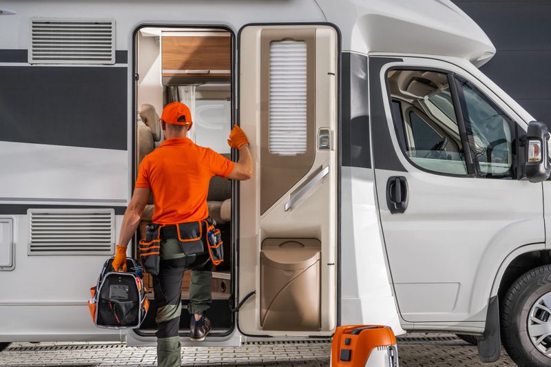 A technician in orange gear approaches a motorhome door, preparing tools for repairs in a modern urban area during daylight hours.
