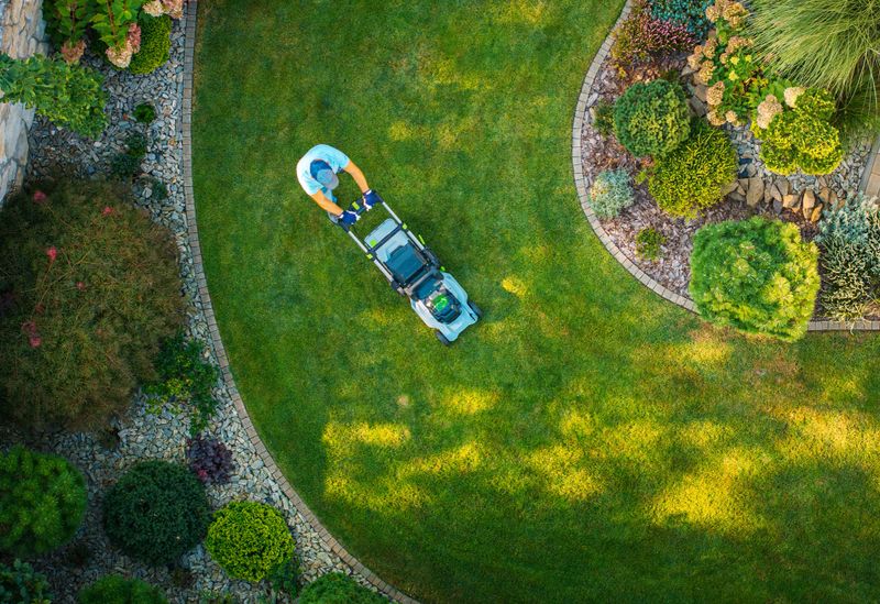 A person mows a lush green lawn in a beautifully landscaped garden with flowerbeds and decorative plants during bright daylight.